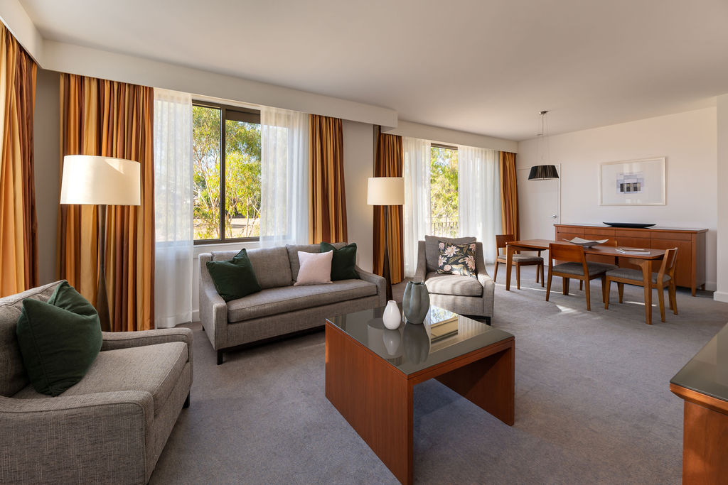 Bright living room with gray sofas, a wooden coffee table, tall curtained windows, and a dining table in the background.