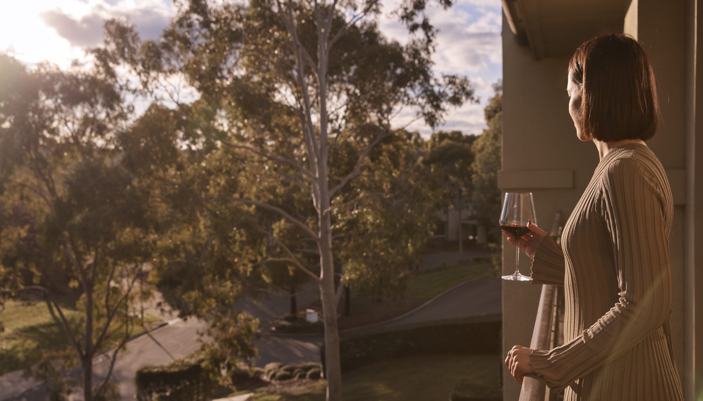 A woman stands on a balcony holding a glass and looking out over a tree-lined landscape in warm evening light.