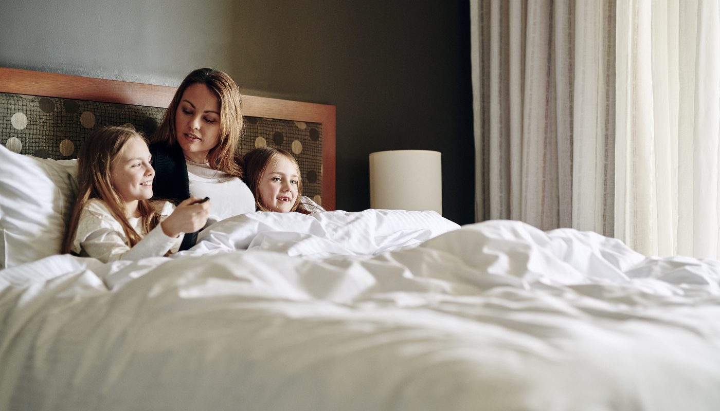 A woman and two young girls sit in bed under white covers, looking at a phone.