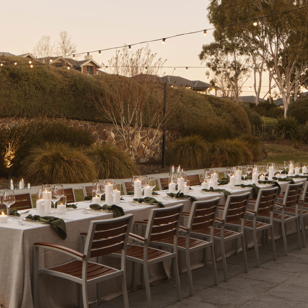 An outdoor dining table set with plates, glasses, and rows of chairs beneath string lights, beside a landscaped garden.