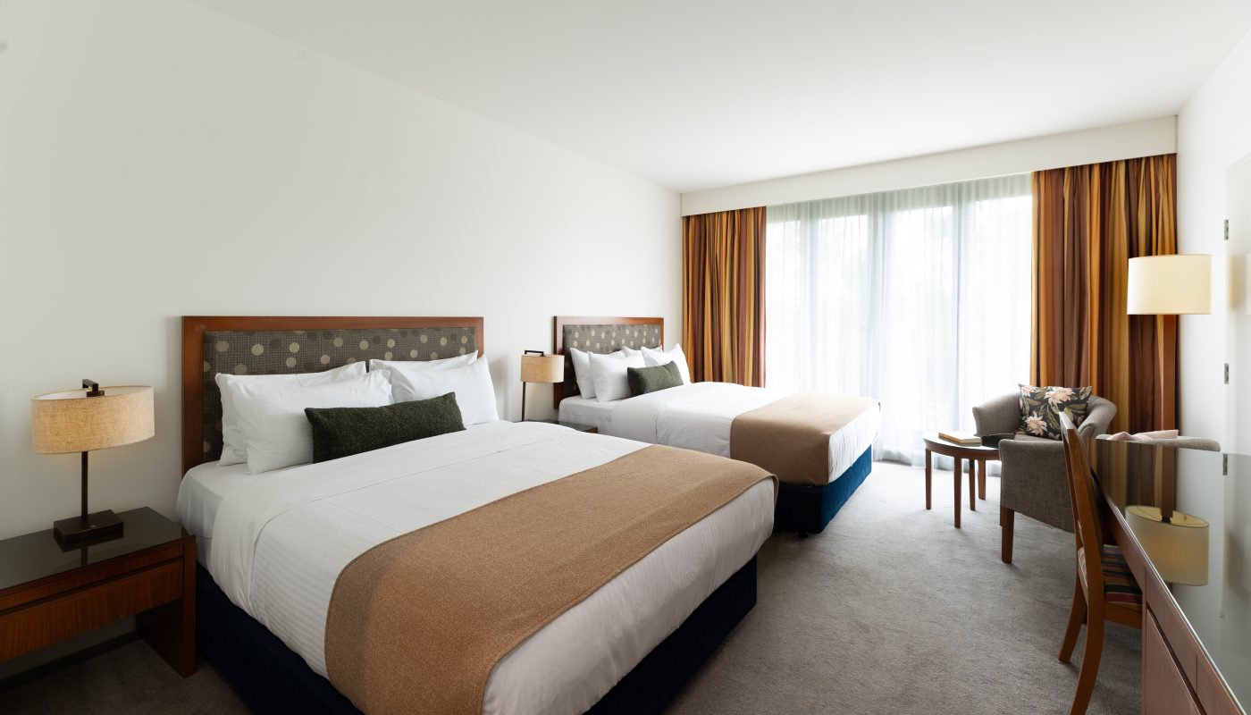 A bright hotel room with two beds, large curtained windows, and a small seating area beside the window.