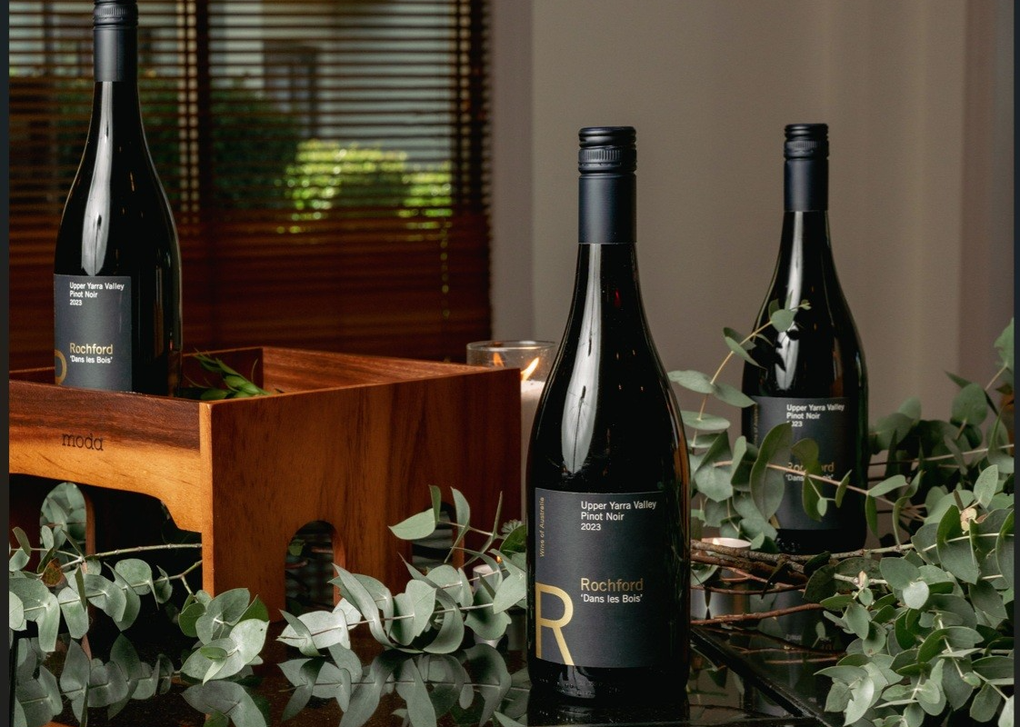 Three dark wine bottles sit on a wooden crate and reflective table, surrounded by leafy branches near a window.
