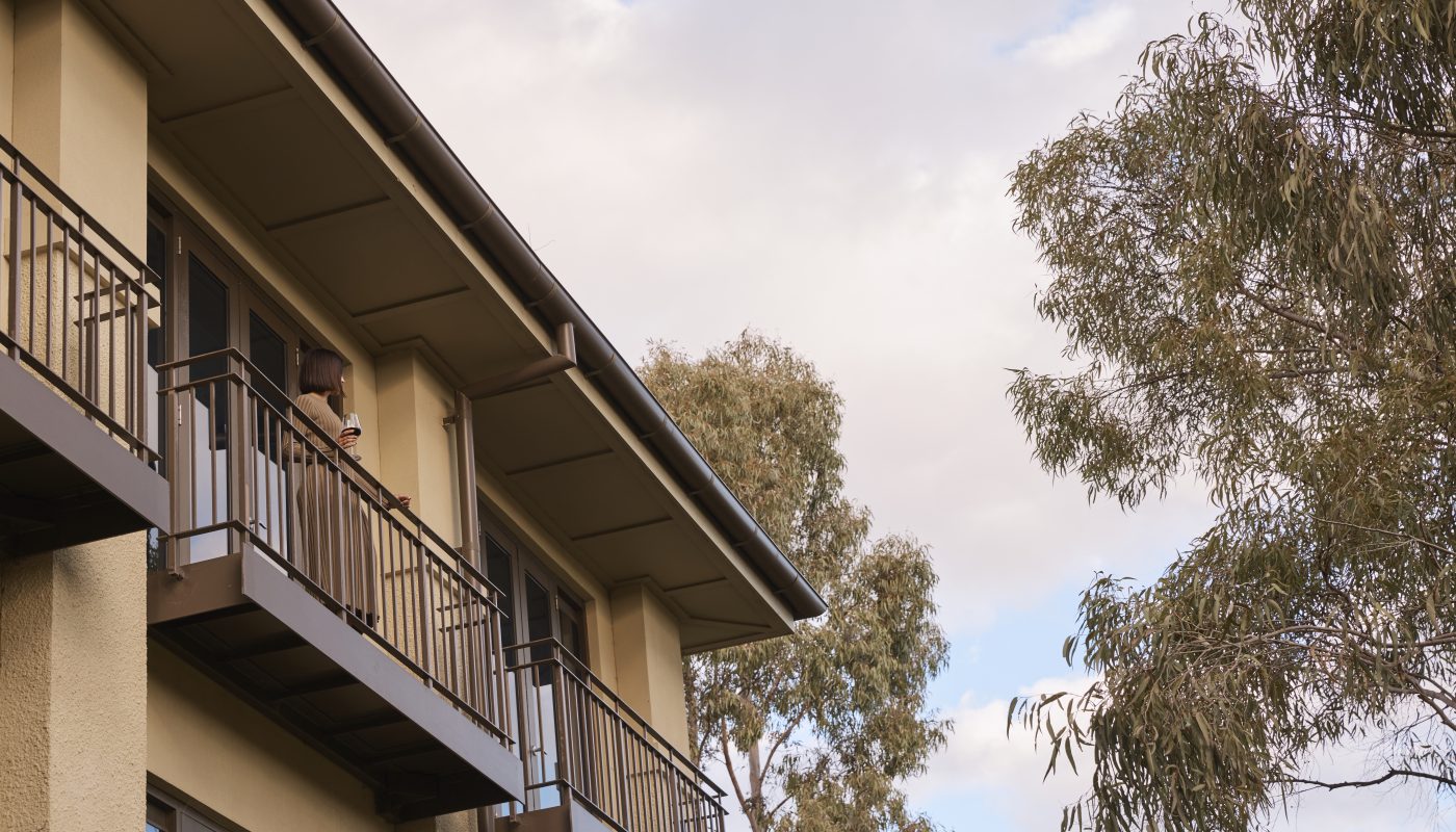 Multi-story apartment building with metal-railed balconies beside leafy trees under a cloudy sky.