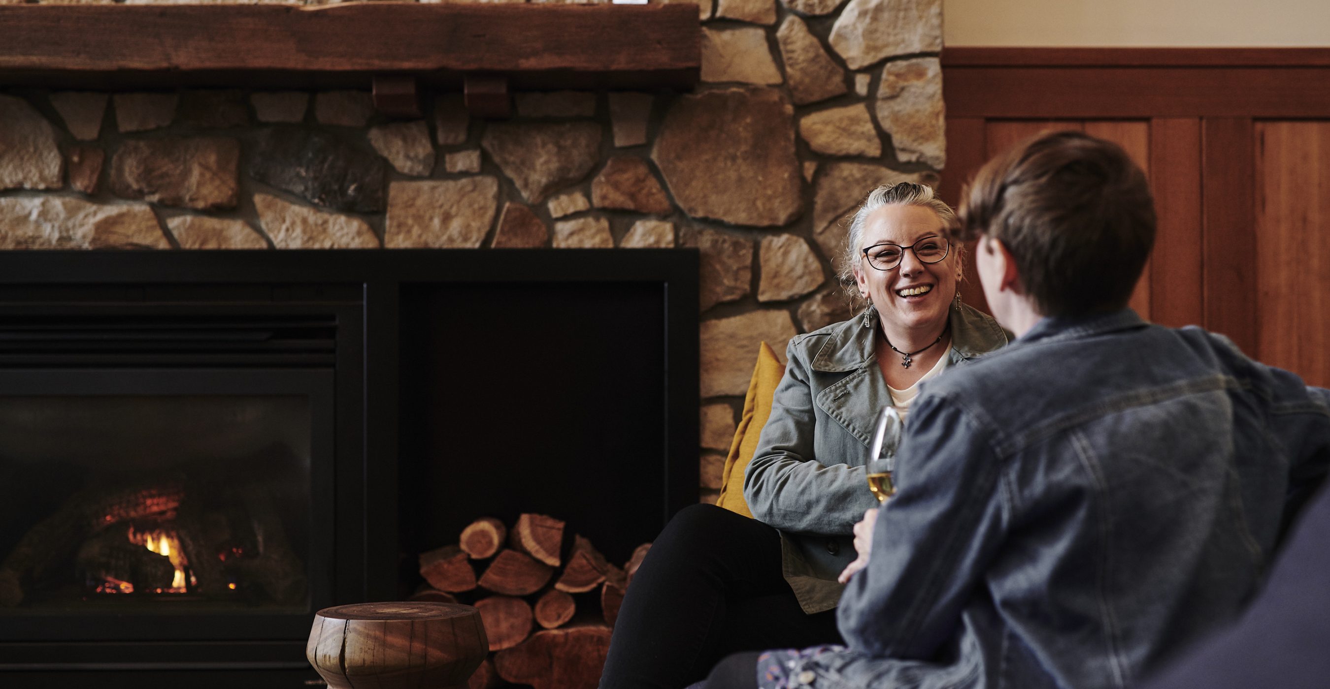 A smiling woman talks with a man seated beside a stone fireplace and wooden mantel.