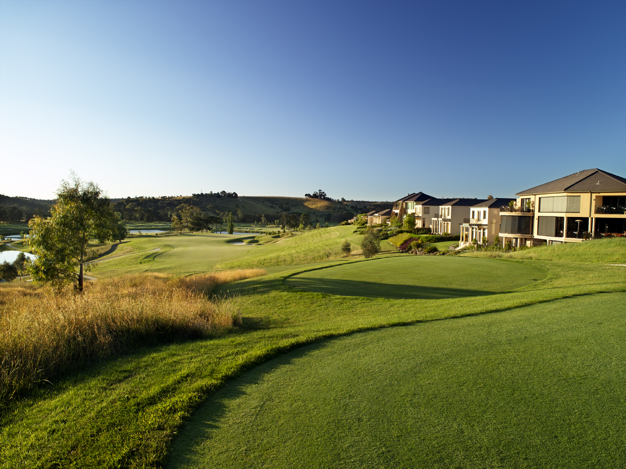 A manicured golf course with rolling green fairways, trees, and houses beneath a clear blue sky.