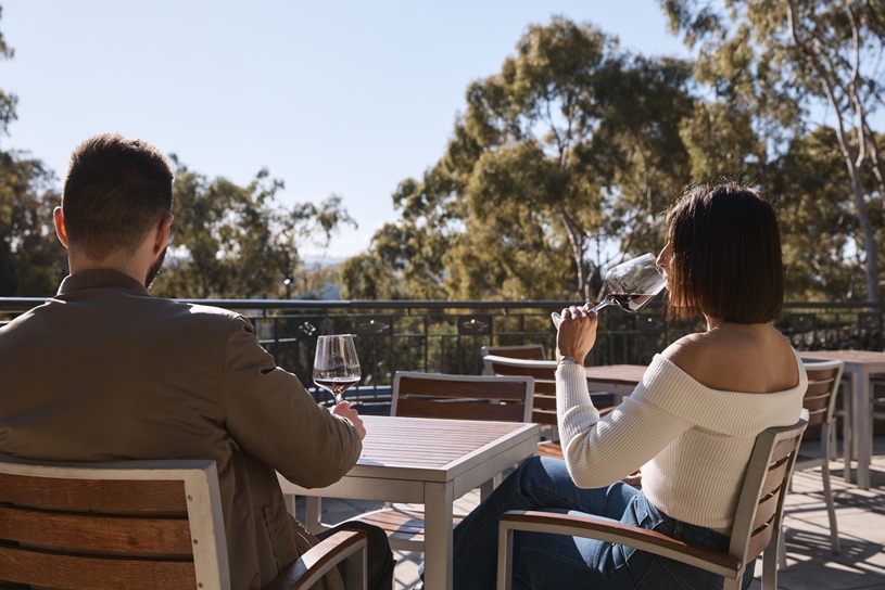 Two people sit at an outdoor patio table, with the woman holding a glass near her face and trees beyond the railing.