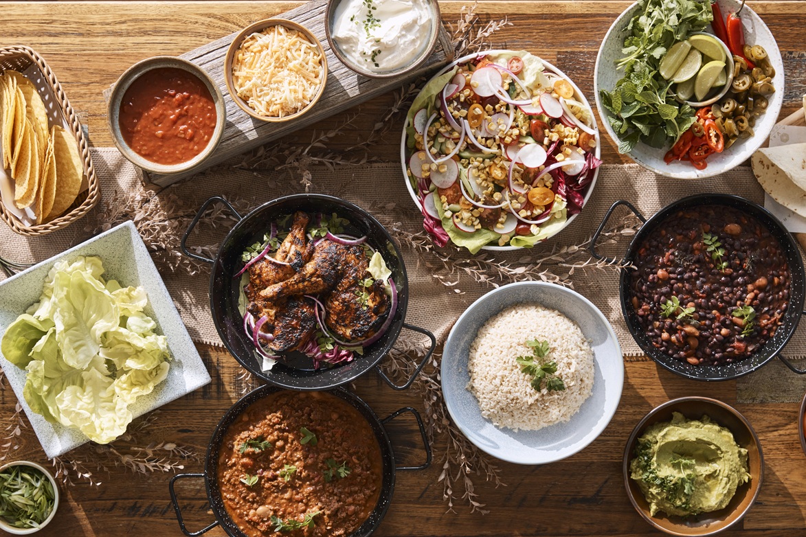 Overhead view of a table spread with bowls of salads, curries, rice, dips, bread, and leafy greens.
