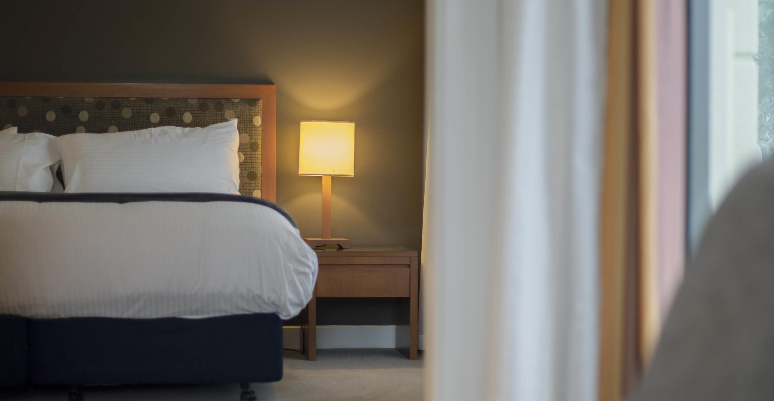 A neatly made bed sits beside a nightstand with a glowing lamp, while curtains cover a window on the right.