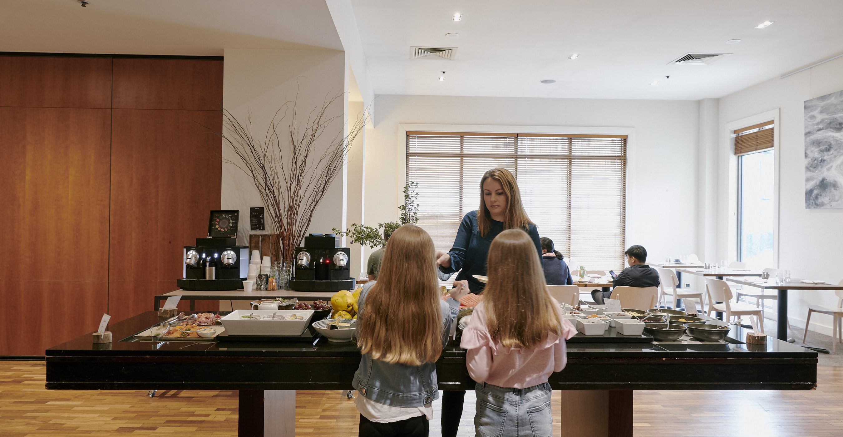 Several people gather around a long kitchen island, with a woman standing behind it and two girls facing her.