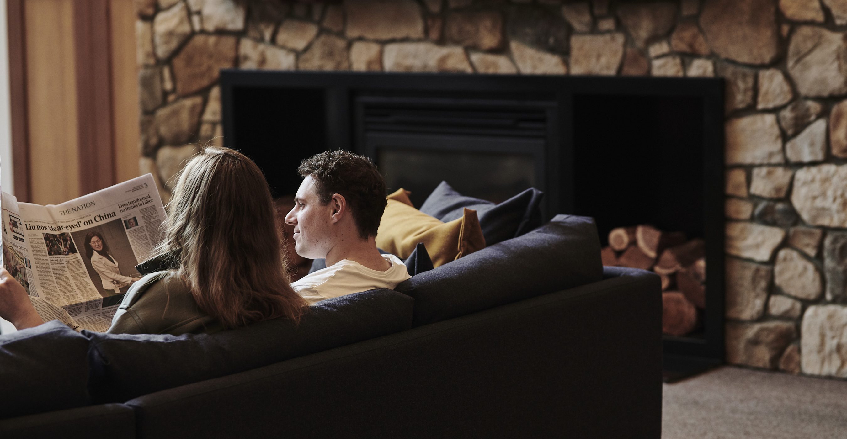 A couple sits on a dark sofa in front of a stone fireplace, with the woman holding a newspaper.