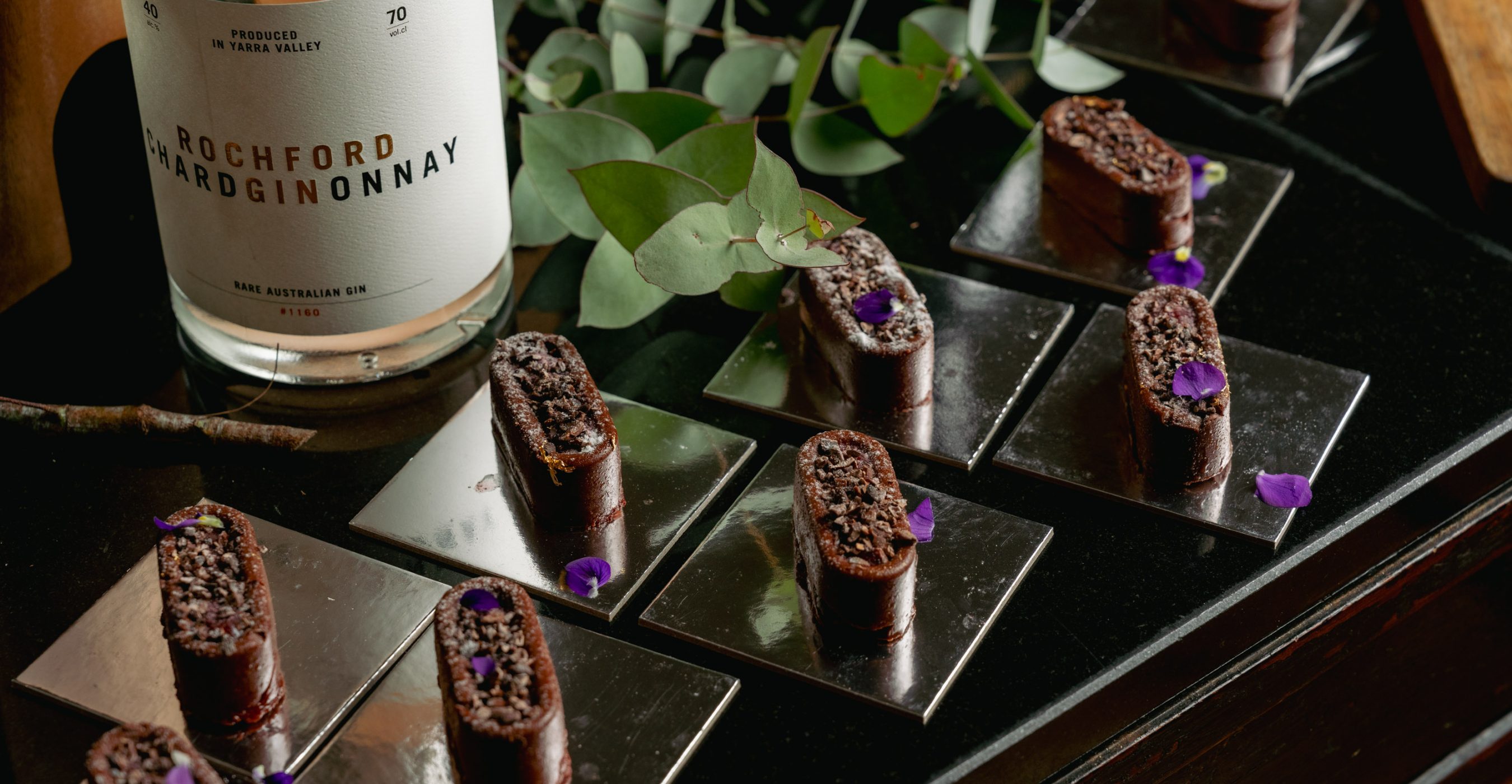 Small chocolate desserts topped with nuts and purple flowers arranged on a mirrored tray beside a bottle.