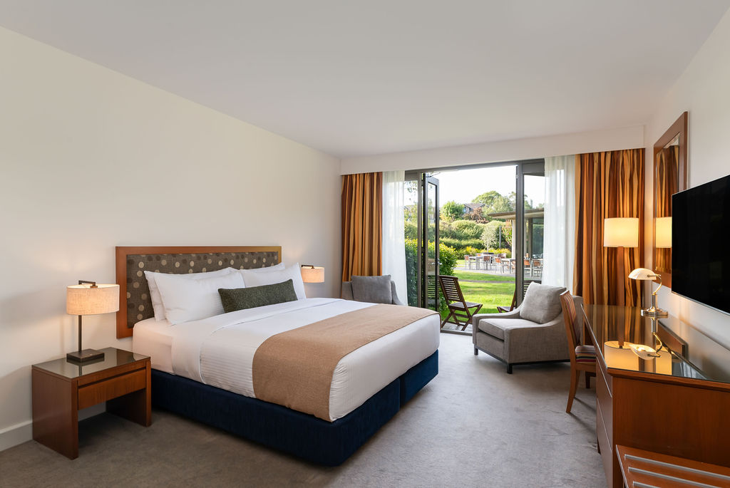 Modern hotel room with a king bed, armchair, and sliding glass doors opening onto a garden.